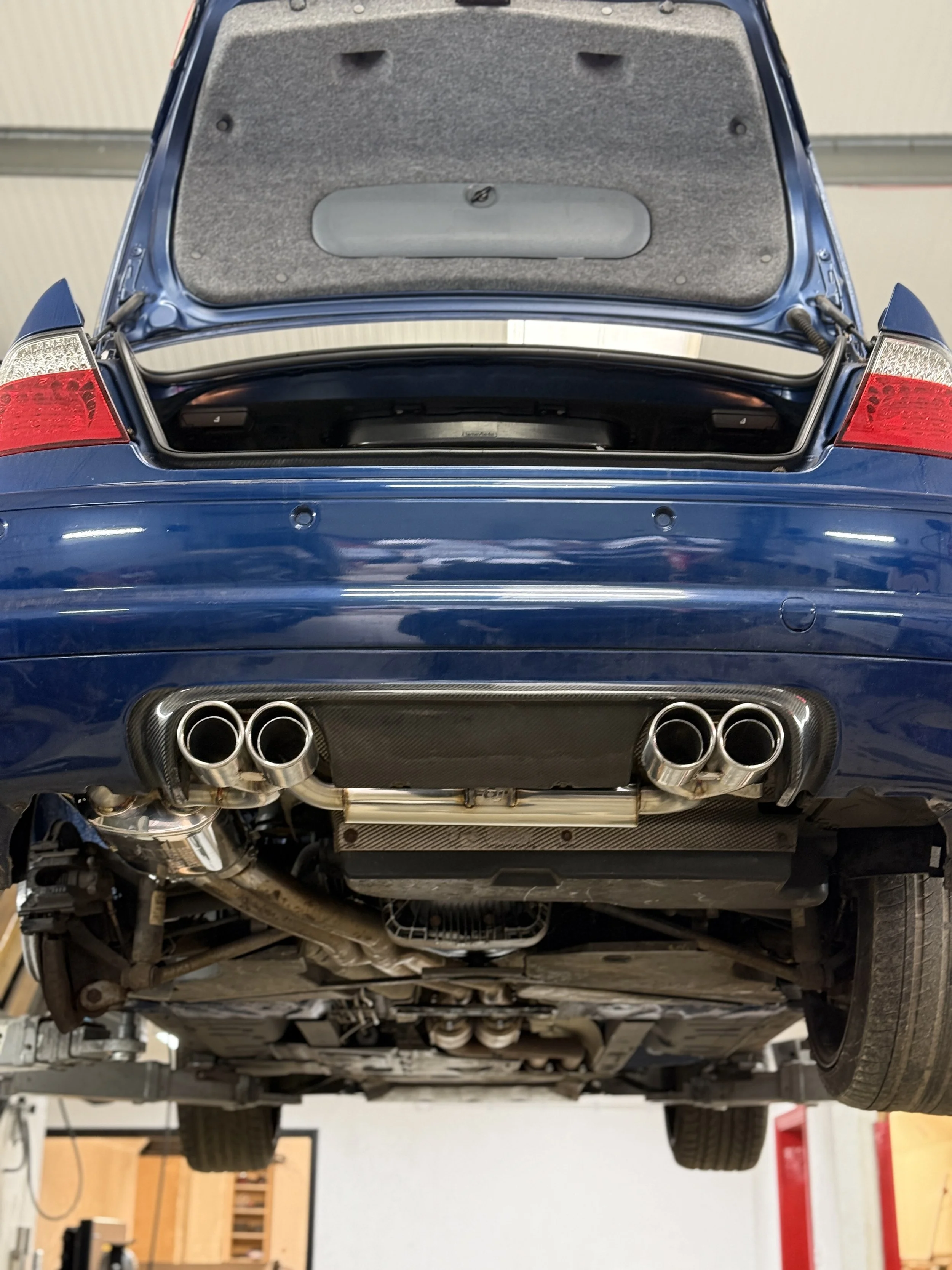 Underneath view of a blue sports car with aftermarket exhaust pipes and visible components in a garage. E46 BMW M3