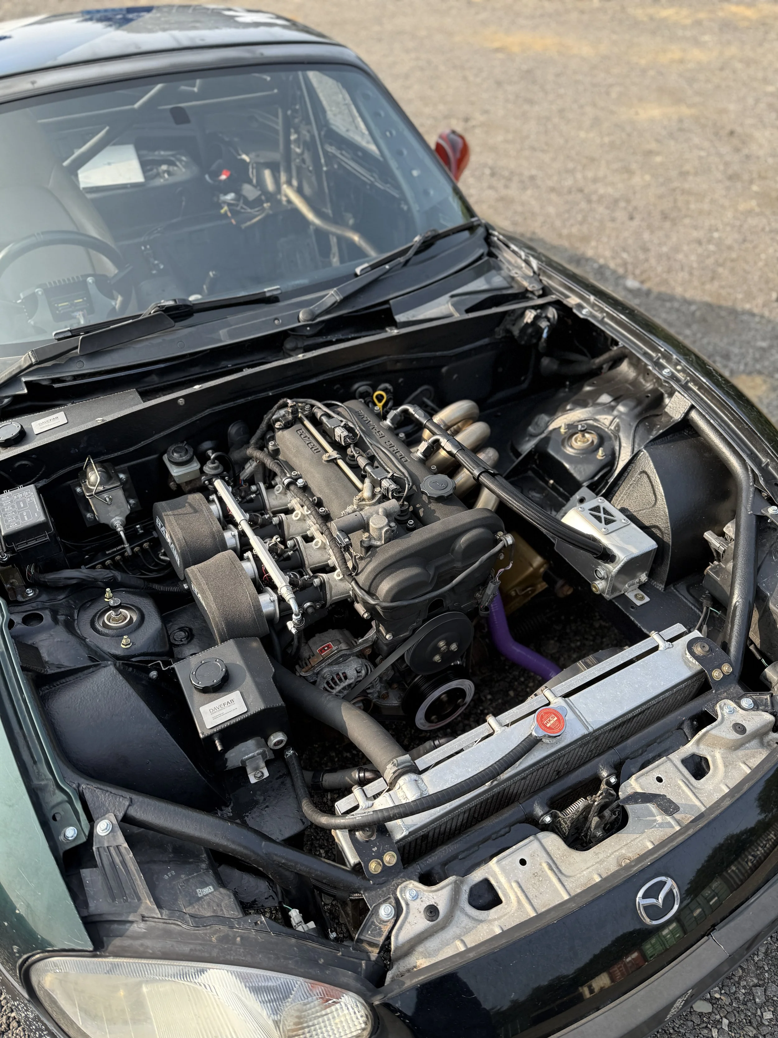 Close-up view of a race car engine inside the front compartment of a black Mazda Mx5 Miata sports car.