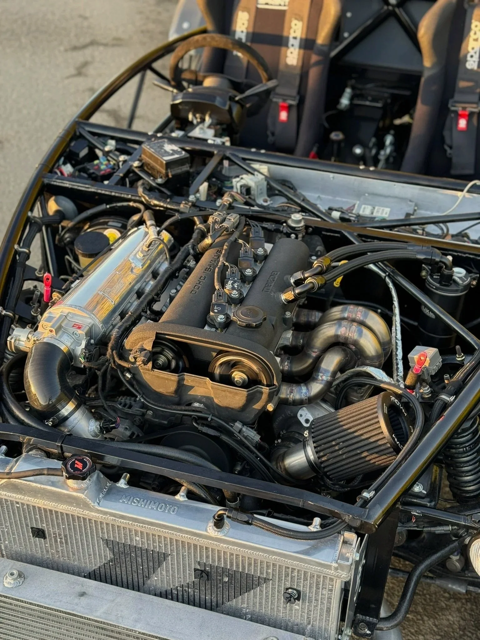 Close-up view of a high-performance car engine with various components, tubes, and wiring, and two racing seats in the background. MEV exocet