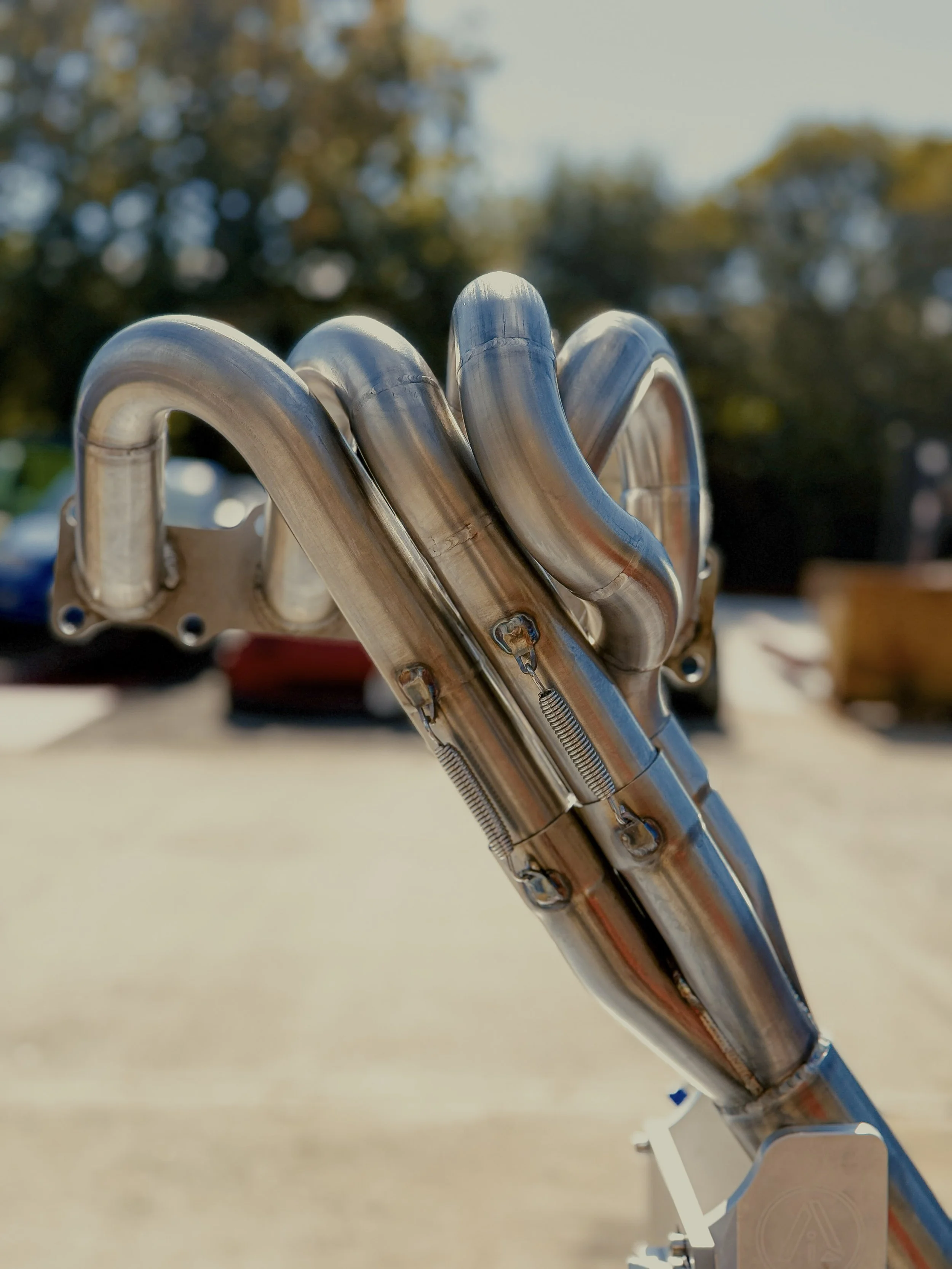 Close-up of a bespoke high rise mx5 miata mazda exhaust manifold, outdoors in a parking lot with blurred trees and cars in the background.