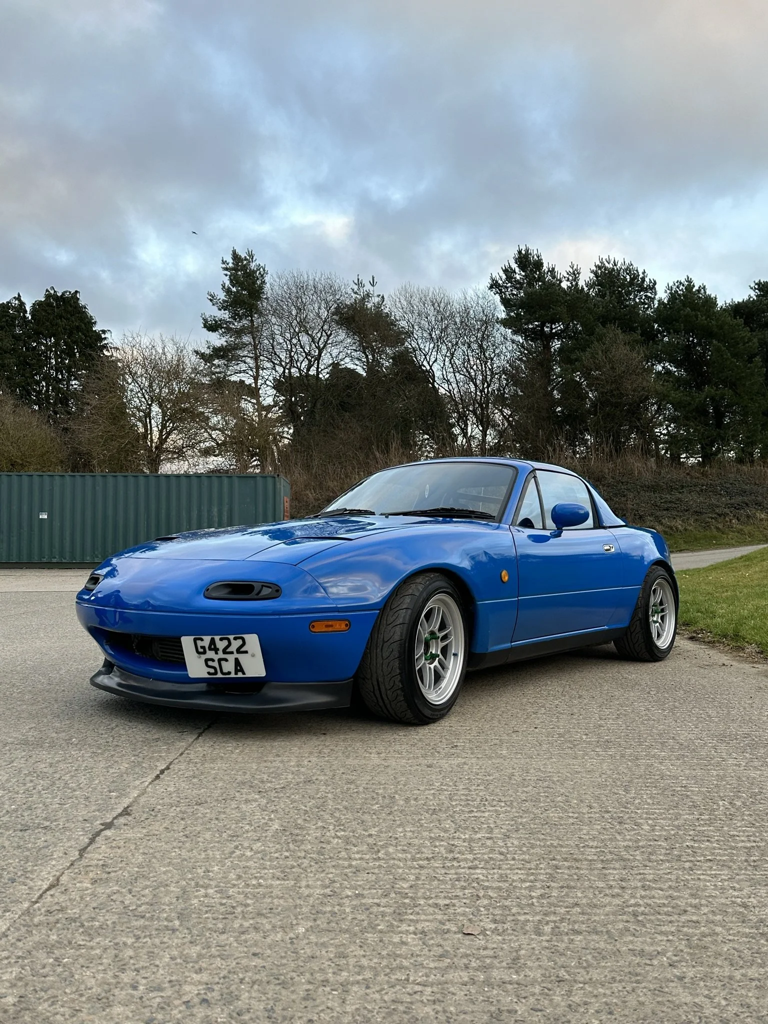 A blue Mazda MX-5 Miata sports car parked on a concrete surface with trees and a green storage container in the background, under a cloudy sky.