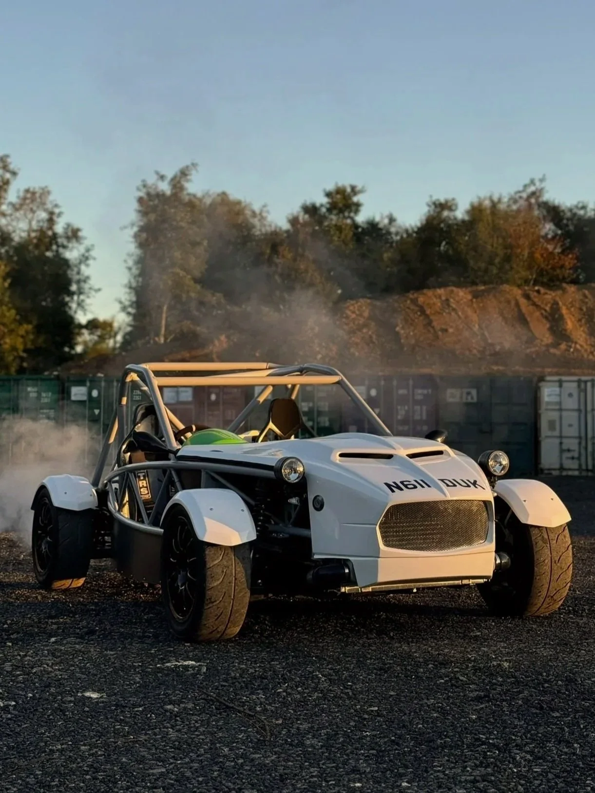 MEV Exocet A white open-cockpit race car with a visible roll cage, parked on a gravel surface with smoke behind it and trees in the background.