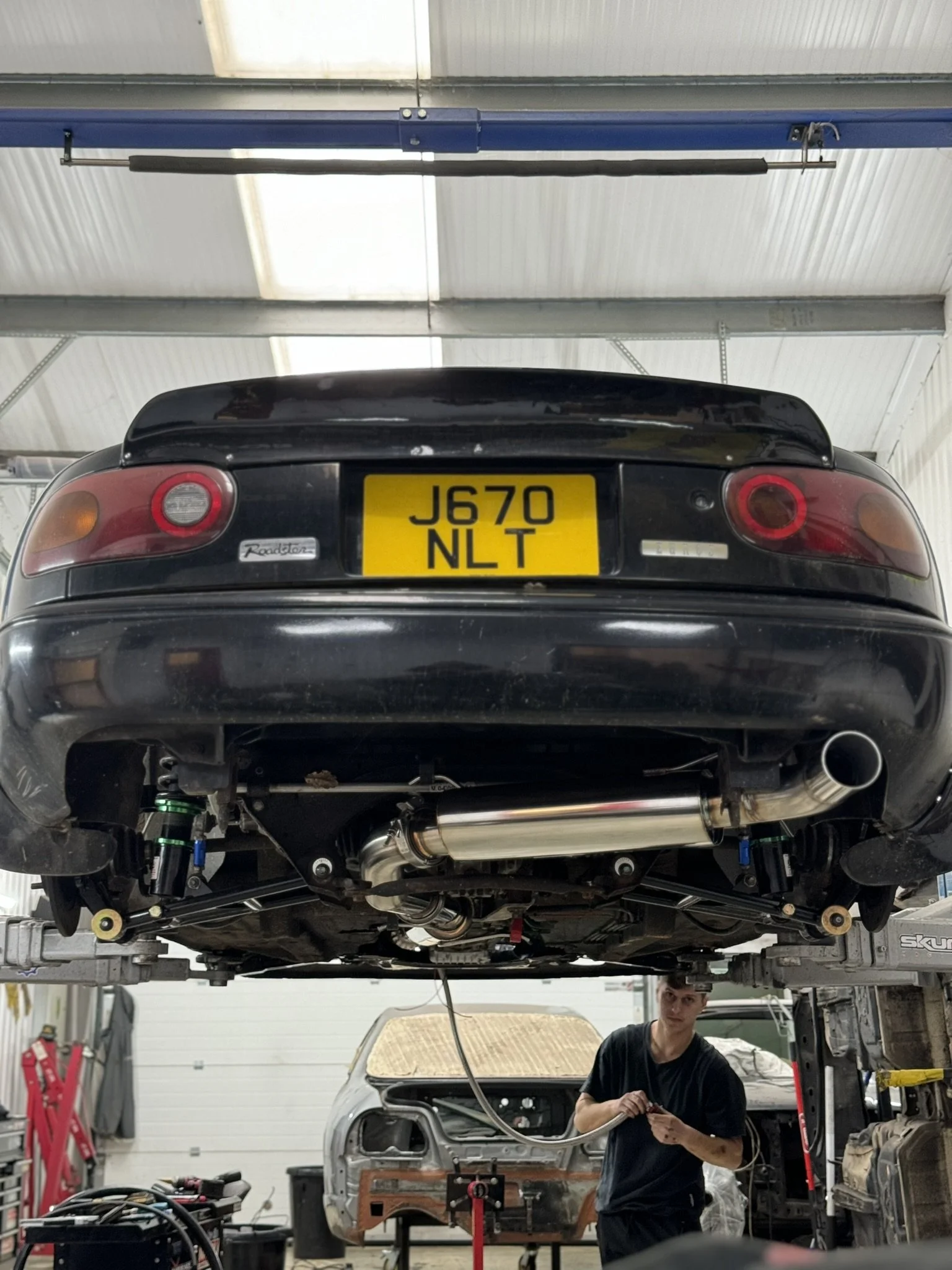 A black Mazda MX-5 Miata sports car lifted on a hydraulic lift in a garage, showing its underside and exhaust system. A mechanic stands below, working.