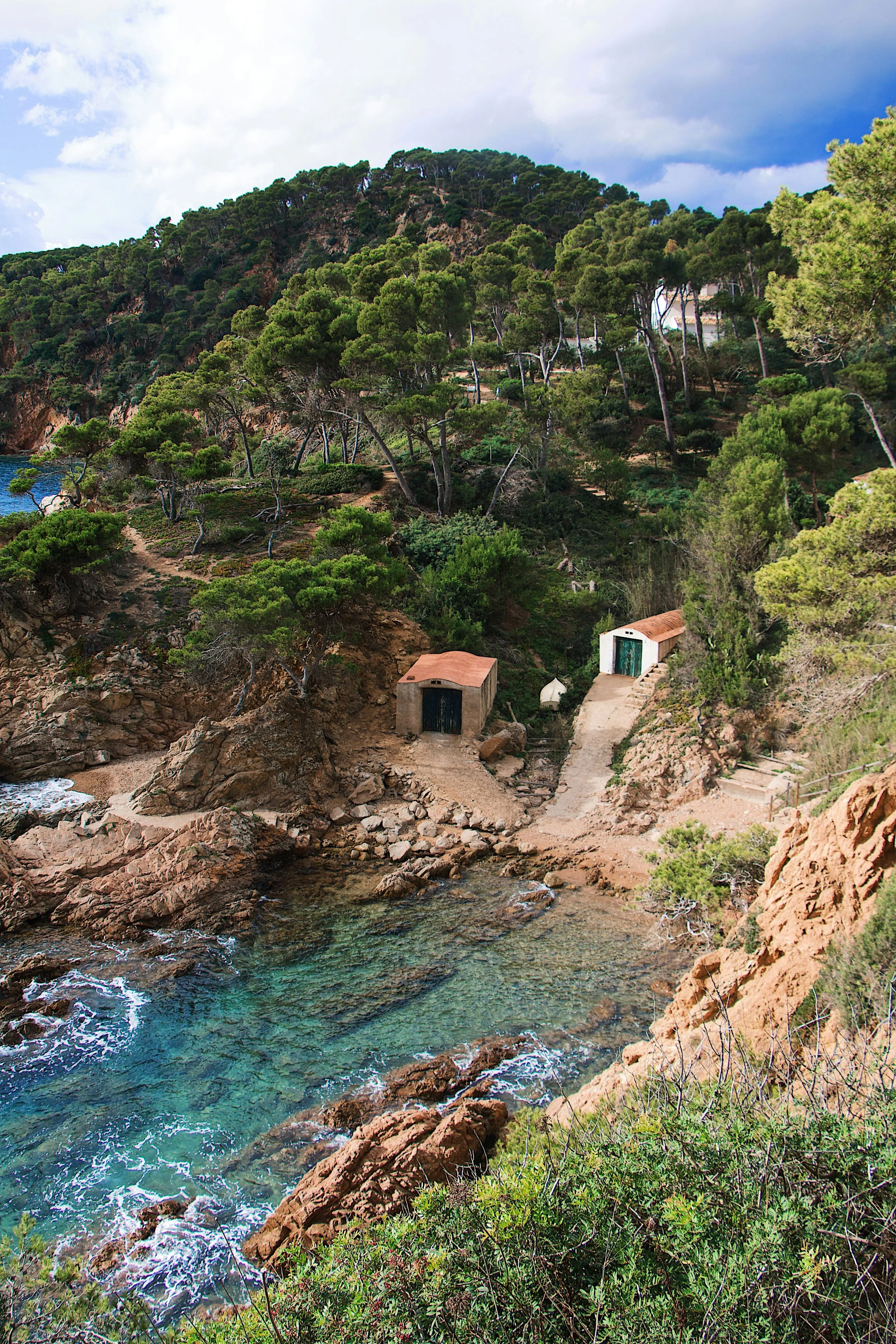 Costa Brava scene with rocky shoreline, clear water, green trees, and two small buildings with red and green roofs.