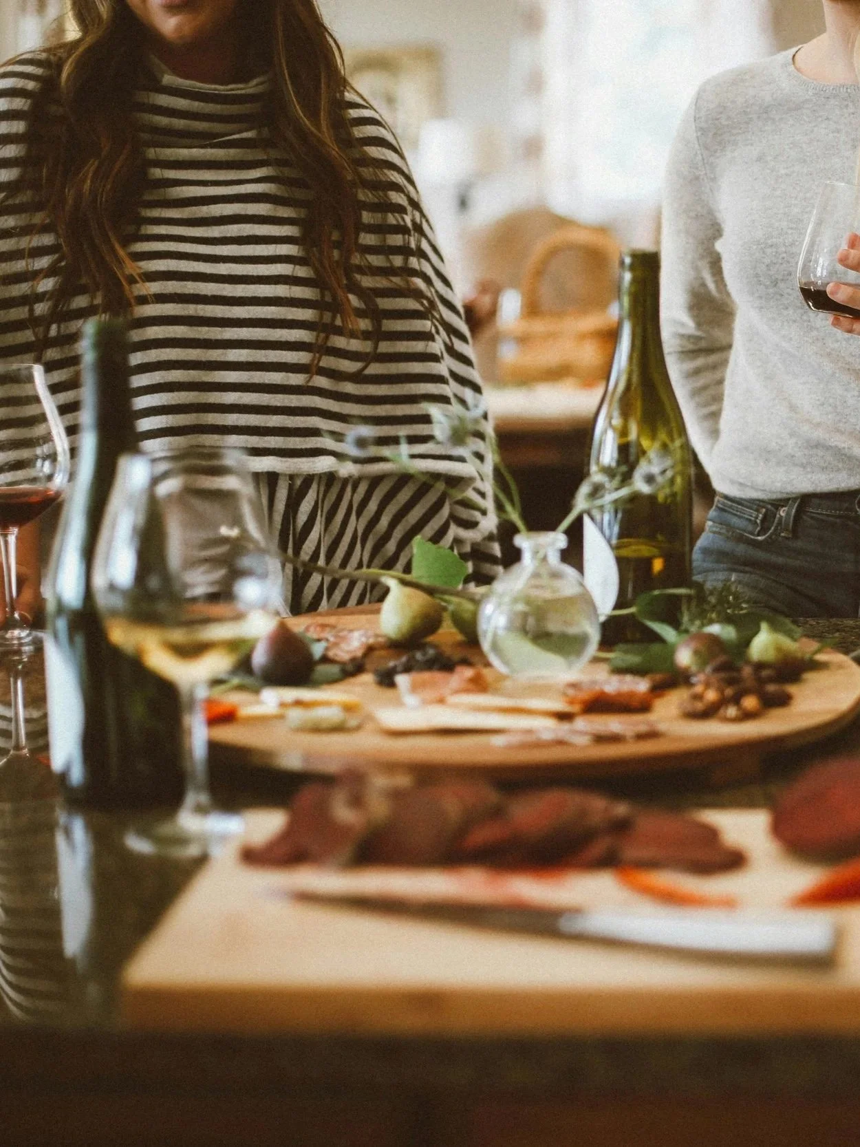 Two women are standing near a dining table with a cheese and charcuterie platter, wine bottles, and glasses, in a cozy home setting.