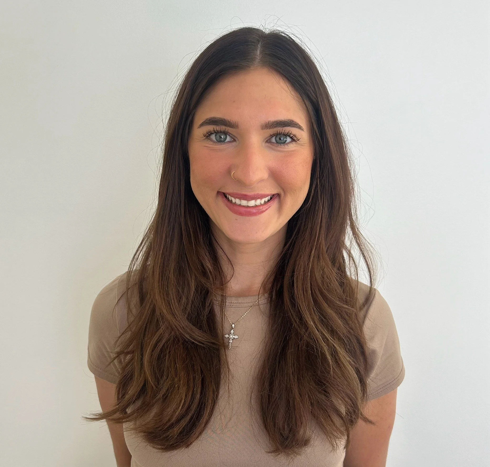 A young woman with long, wavy brown hair, blue eyes, wearing a beige t-shirt, a necklace with a cross pendant, and a nose ring, smiling in front of a plain white wall.