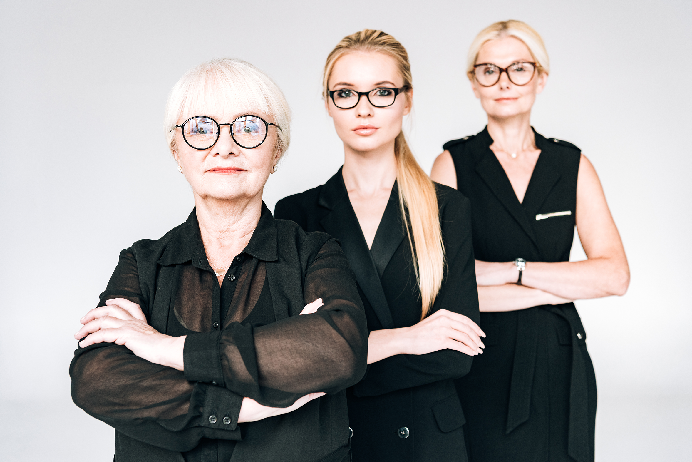 Three professional women standing with arms crossed, wearing black business attire and glasses, looking confidently at the camera against a plain white background.