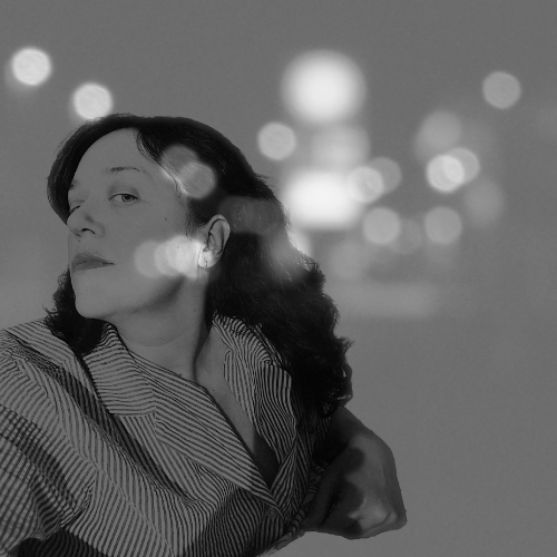 A black and white photo of a woman with dark hair and striped shirt, looking over her shoulder, with blurred lights in the background. Promotional photograph of acoustic dark folk artist, crowspeak.