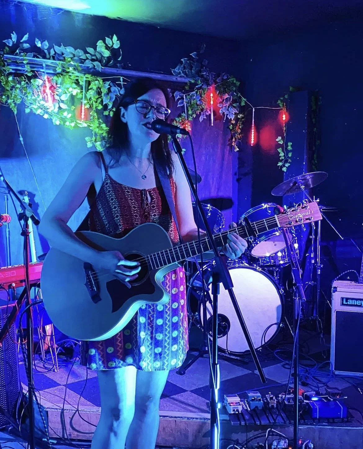 A woman is singing into a microphone while playing an acoustic guitar on a stage with blue and purple lighting and decorative greenery. crowspeak performing live with a tangle wood acoustic guitar and singing.