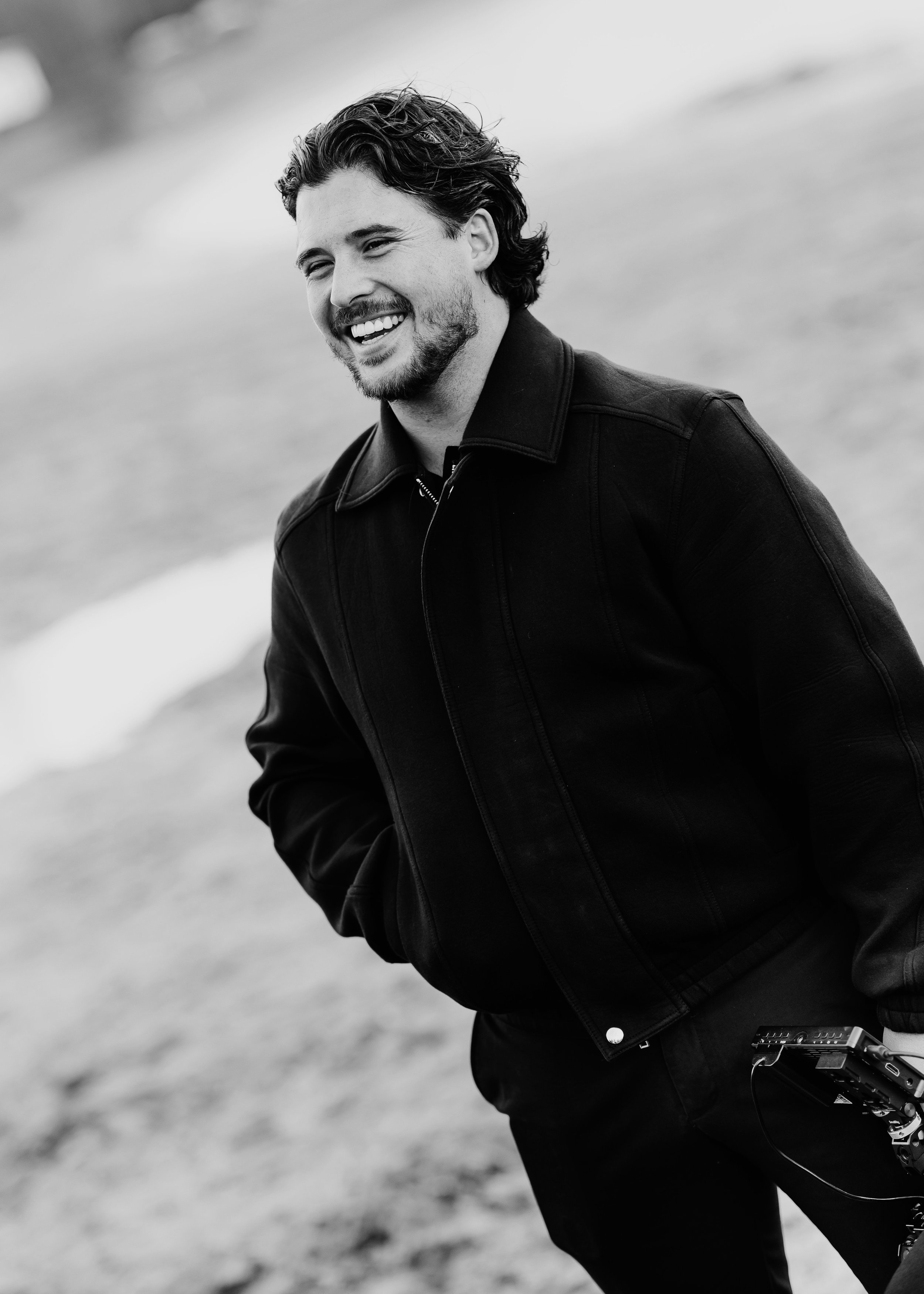 Een lachende man met warrig haar en een baard, gekleed in een zwarte jas, staat buiten op een natuurlijke omgeving.