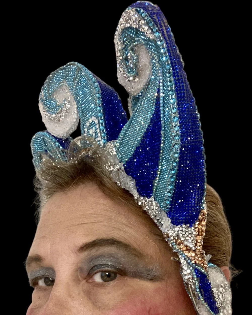Close-up of a woman wearing an elaborate, bejeweled blue and white butterfly-shaped headpiece with intricate patterns and rhinestones against a black background.