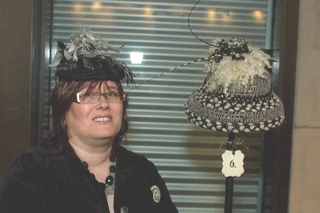 Woman wearing a fancy hat with floral decorations, standing in front of a window with blinds and a matching hat on a display stand.