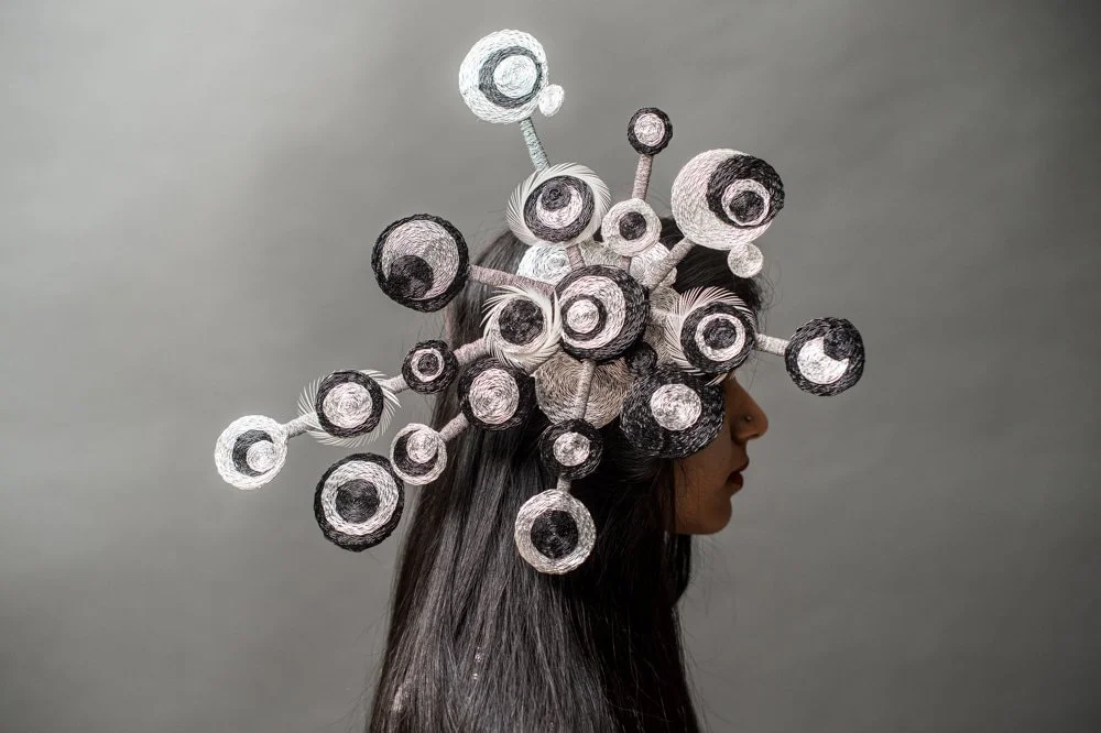Side view of a woman with long black hair wearing an avant-garde headpiece made of numerous interconnected circular elements in black, white, and silver colors.