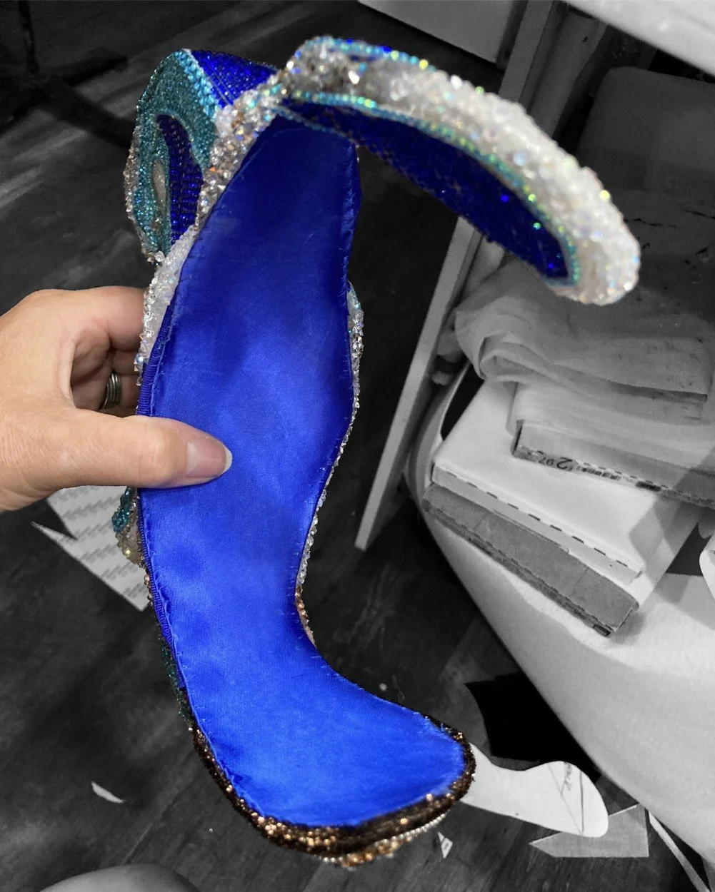 A person's hand holding a shiny, decorated blue and black shoe with a sparkly border, likely a costume or fancy shoe, indoors near a small table and papers.