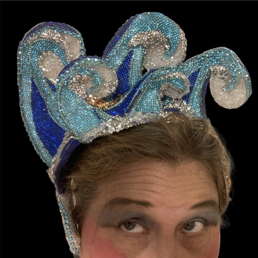 A person wearing an elaborate, sparkly headdress with swirling blue and silver designs and rhinestones against a black background.