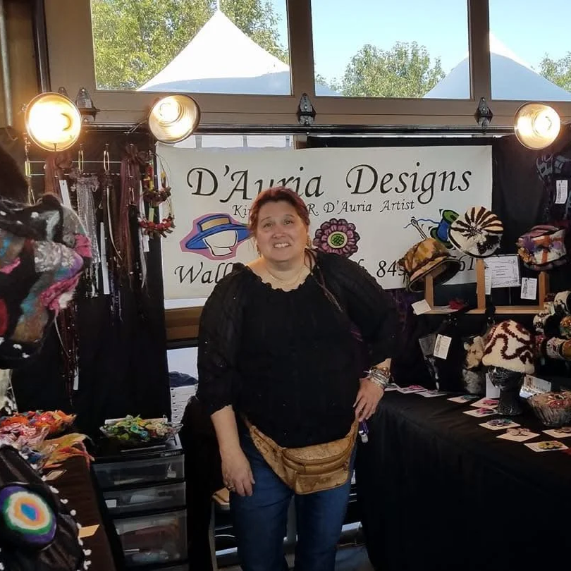Woman standing behind a display table at a craft fair, with jewelry and crafts. A banner behind her reads "D'Auria Designs" with a subtitle "Kid's Art, Jewelry, & D'Auria Artist."
