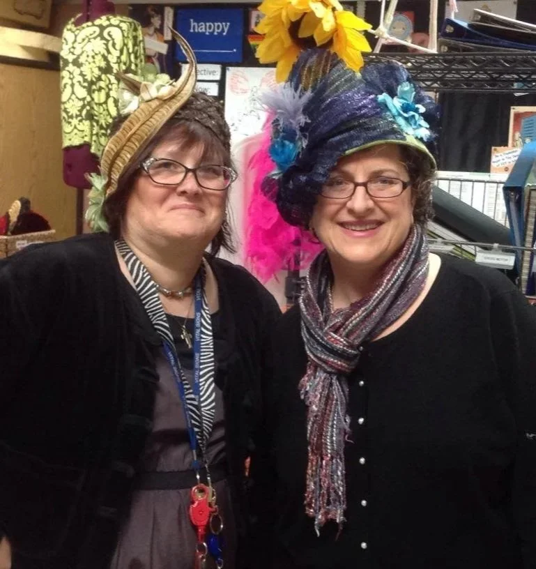 Two women wearing colorful, elaborate hats and glasses, smiling indoors.