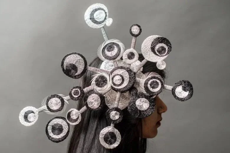 A person with long hair facing sideways, wearing an intricate headpiece made of interconnected spherical eye structures.