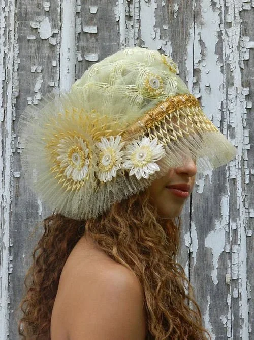 Woman with curly hair wearing a large, decorative hat made of yellow and white fabric and lace, adorned with flowers and sequins, in front of a weathered wooden background.