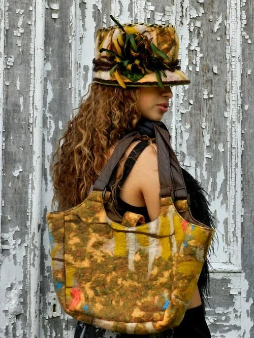 A woman with long curly hair wearing a decorative hat with flowers and feathers, a black sleeveless top, and carrying a large, colorful, patterned tote bag, standing in front of a weathered, peeling gray wooden wall.