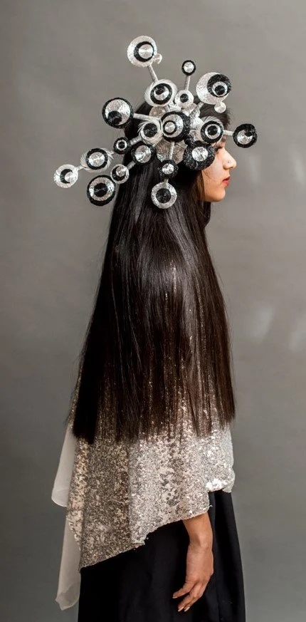 Woman in a silver, sequined top with long black hair and an elaborate metallic headpiece resembling molecules.