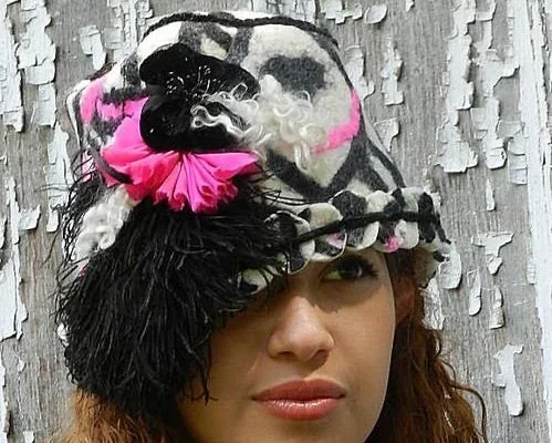 A woman wearing a colorful, artistic hat with a skull design, pink accents, and feather details, standing in front of a textured wooden wall.