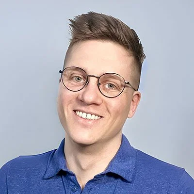 A young man with glasses and short brown hair, smiling, wearing a blue collared shirt, against a light gray background.