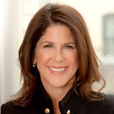 Close-up of a smiling woman with shoulder-length brown hair and blue eyes, wearing a black blazer, indoors with a blurred background.