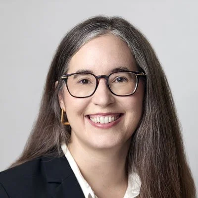 A woman with long brown hair, glasses, and a big smile, wearing a black blazer and white shirt, posing against a plain light gray background.