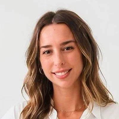 A woman with long, wavy brown hair and light skin, smiling at the camera, wearing a white top.