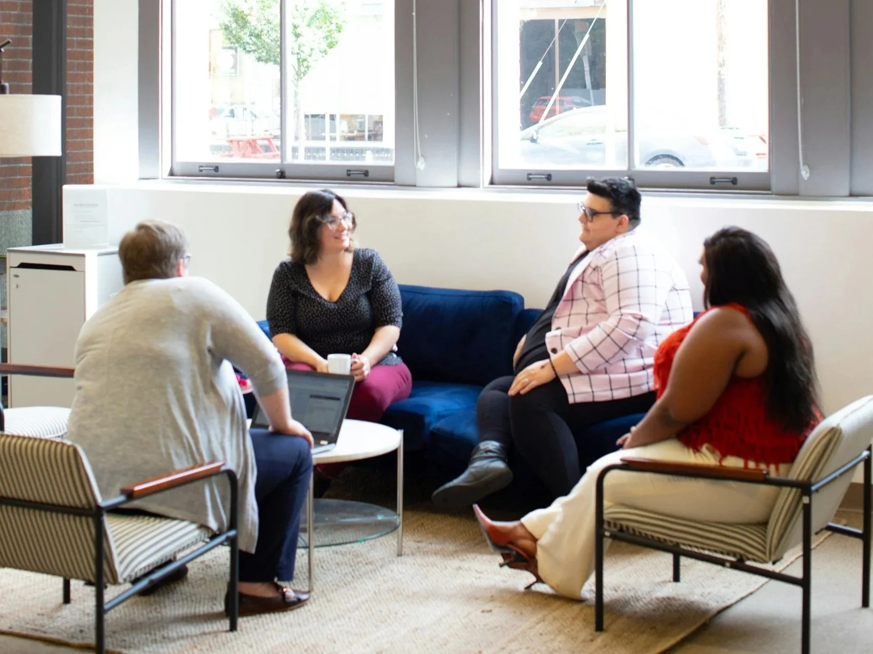 Five people sitting in a casual meeting room, engaged in conversation. Two women and one man are sitting on a blue couch, while another woman with a notebook and a person with a laptop are seated on striped armchairs. Large windows behind them provide natural light.
