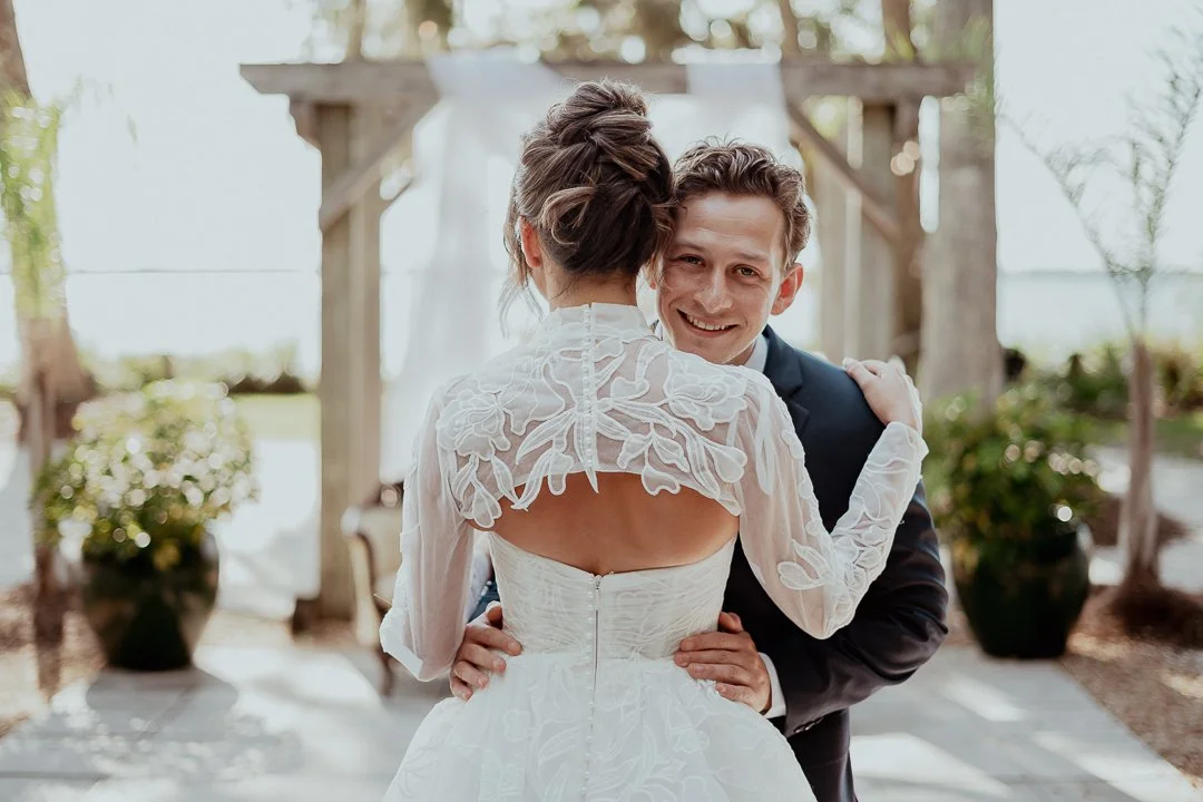 A quiet, intimate moment captured in soft, natural light. The bride stands facing the ocean, her elegant updo revealing the graceful lines of her neck and shoulders. Over her right shoulder, the groom leans in gently — his smile warm, relaxed, and fu