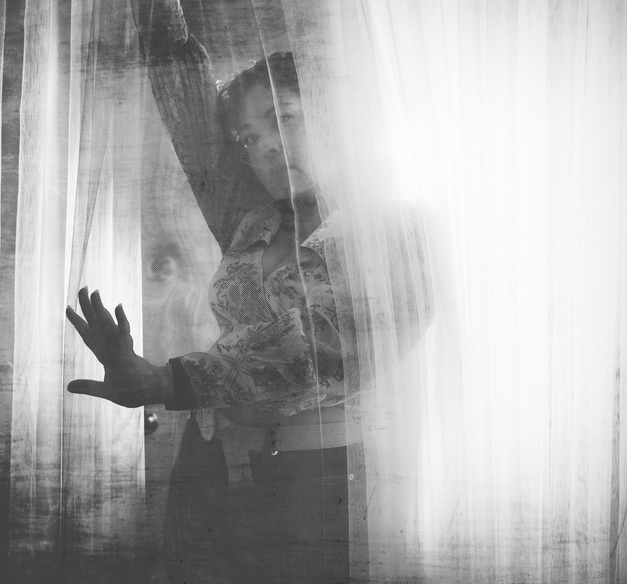Autorretrato en blanco y negro de una mujer detrás de una cortina, sosteniéndola suavemente mientras la luz entra.