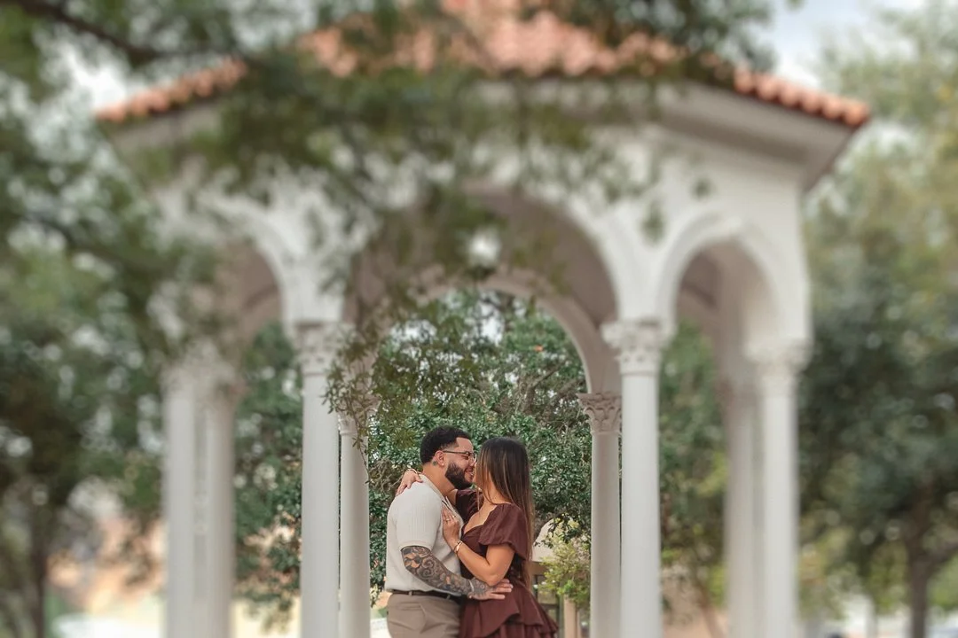 A couple embracing under a white, arched gazebo with trees in the background.