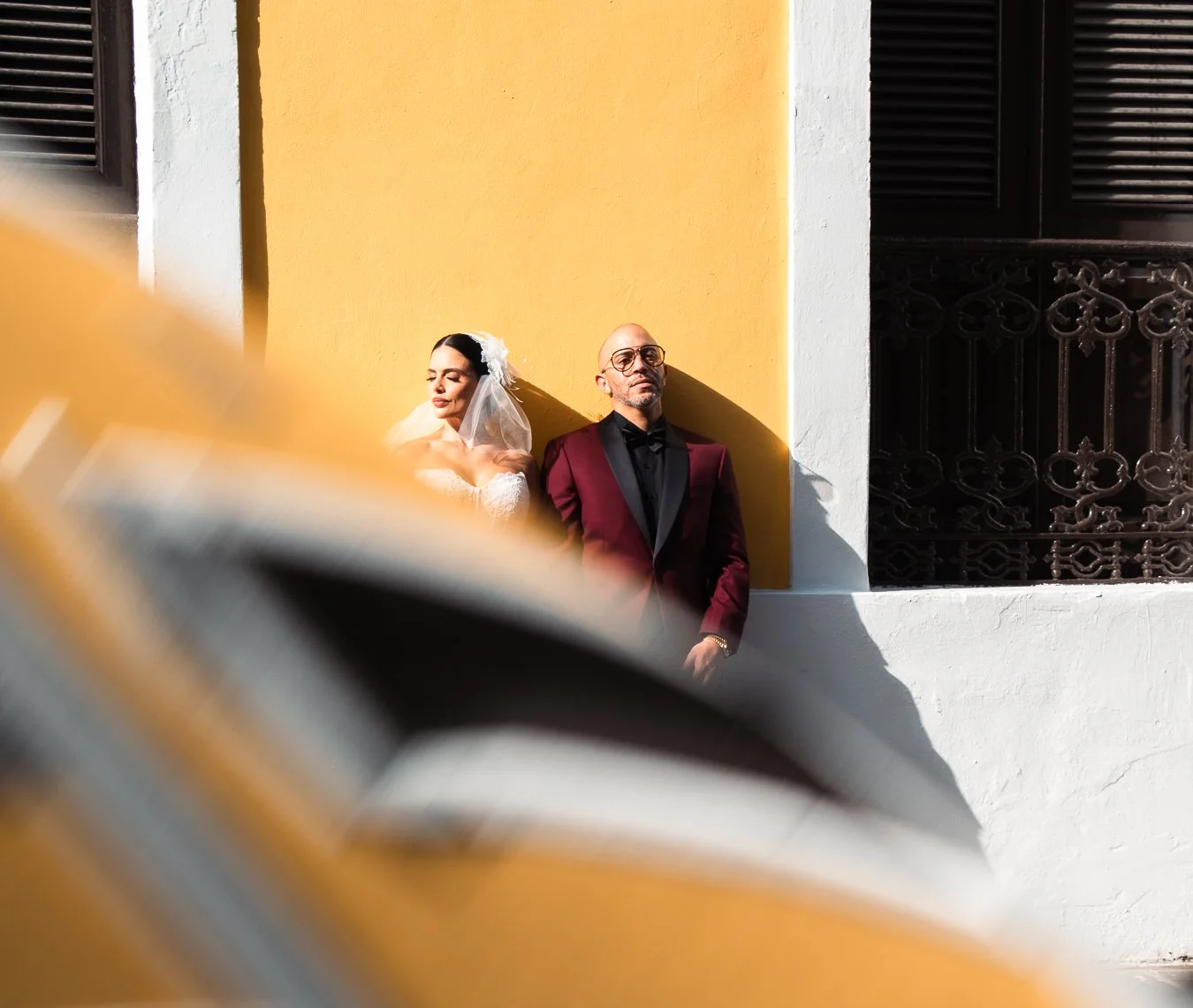 A bride and groom standing against a yellow and white wall with black window shutters, leaning on the wall with their eyes closed in bright sunlight.