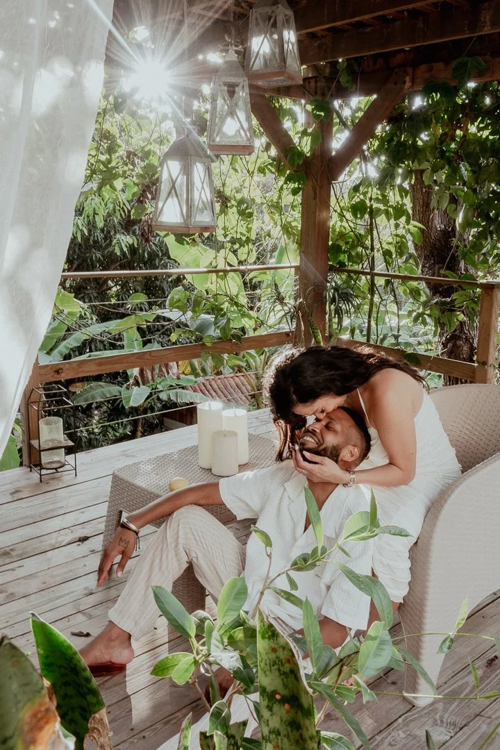 A woman and a man sharing a tender moment on a wooden outdoor deck surrounded by greenery and sunlight, with lanterns and candles nearby.