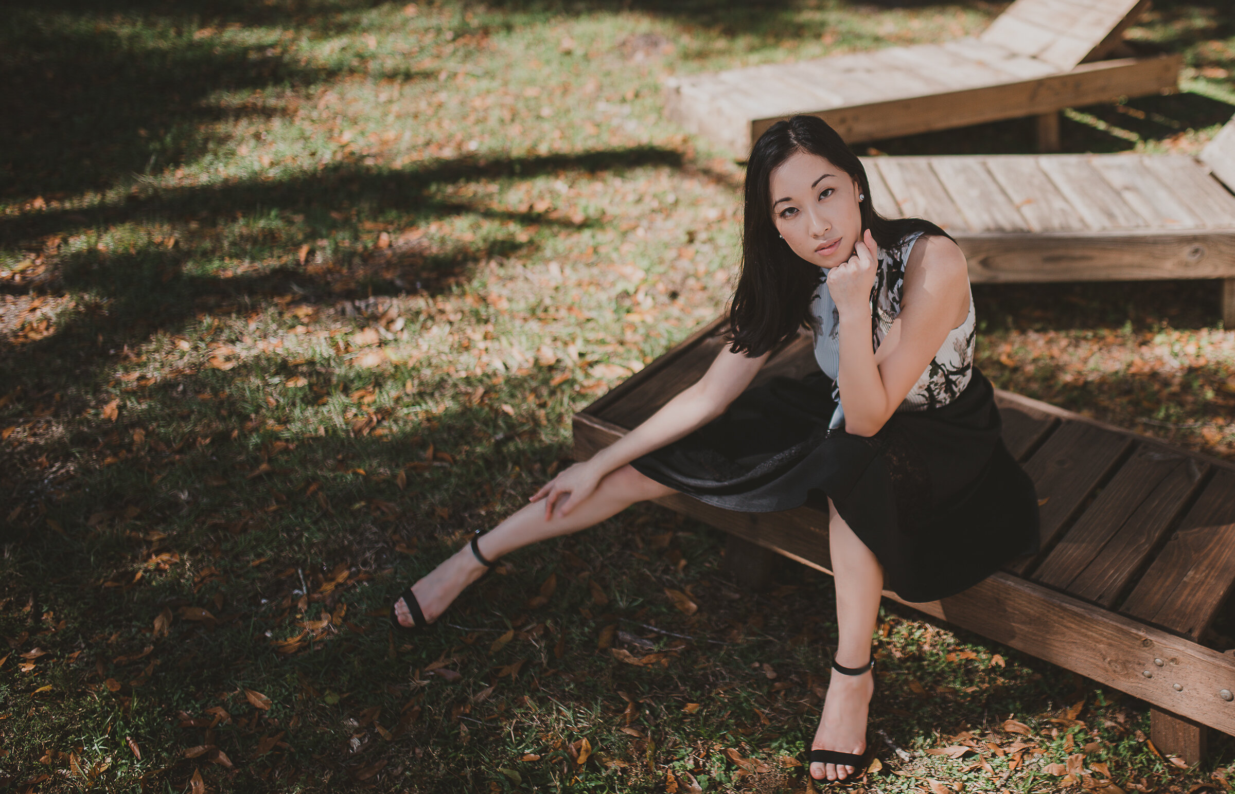 A young woman in a black dress with lace details sitting on a wooden platform outdoors, with one leg extended and the other bent, surrounded by fallen leaves and shaded by trees.