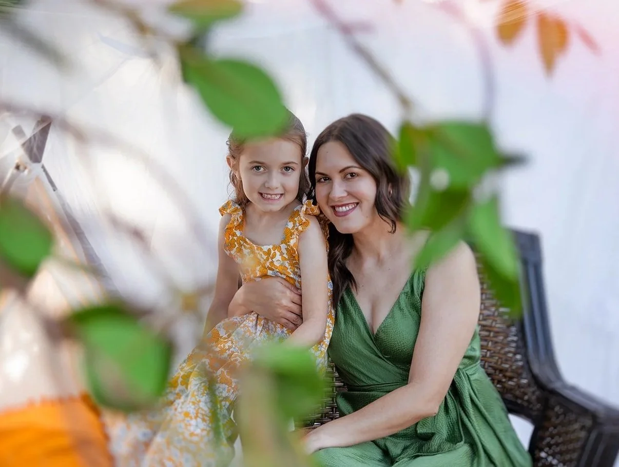 A smiling woman and a girl sitting on a wicker chair, framed by green leaves in the foreground.
