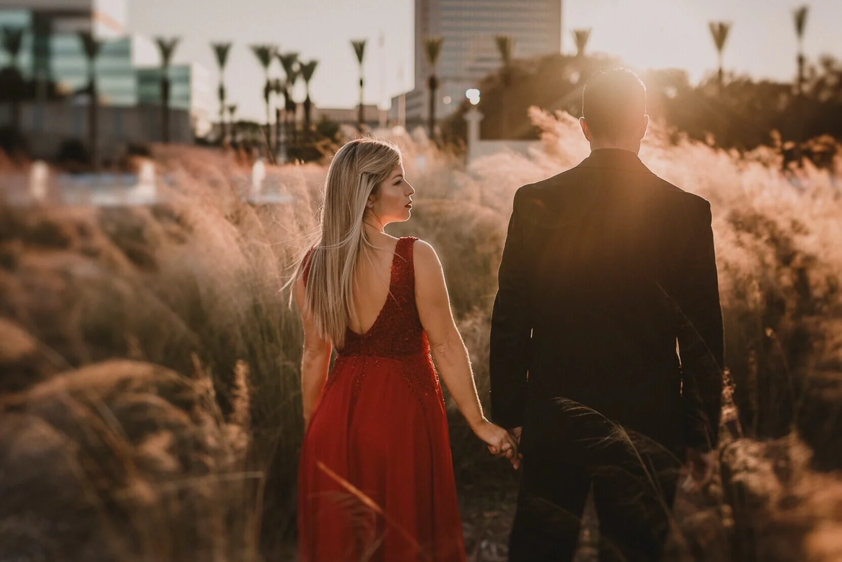 A couple holding hands walking through a field with tall grass during sunset, with a cityscape and palm trees in the background.