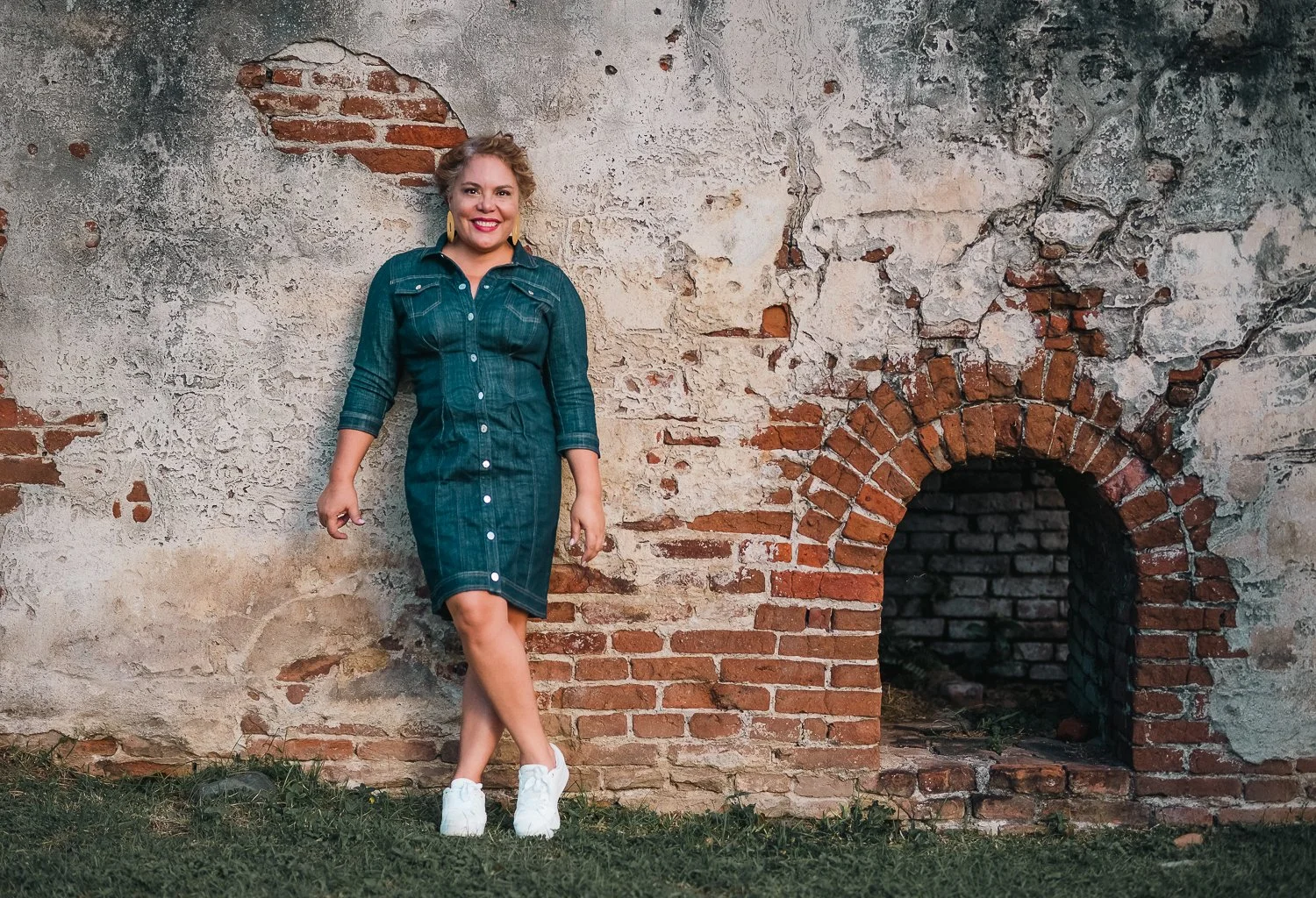 A woman with curly blonde hair wearing a denim dress and white sneakers stands against a weathered brick and stucco wall, smiling at the camera. Karen Fabiola Photo