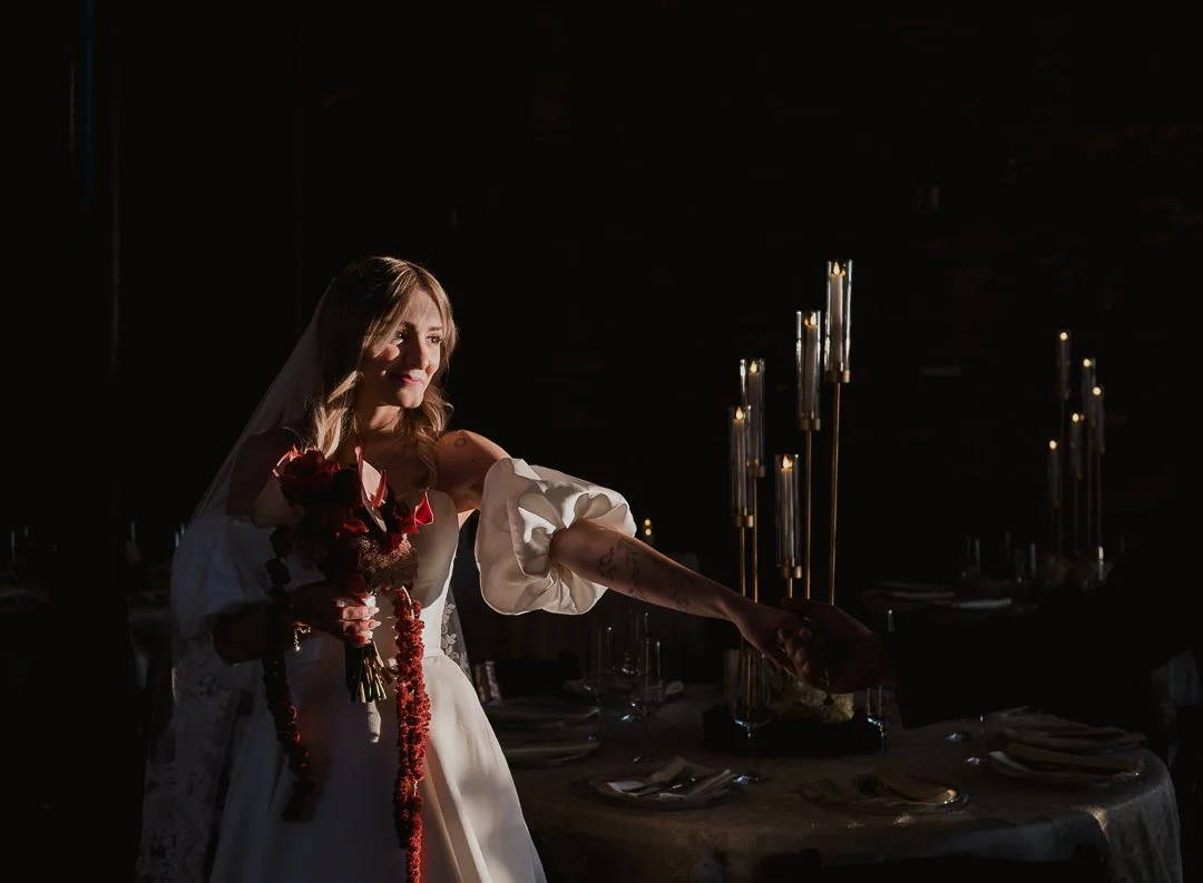 A romantic, dark and moody wedding photograph capturing a bride reaching for the groom’s hand during an intimate moment at their wedding venue. The image highlights a cinematic connection between the couple, with soft shadows, rich tones, and elegant
