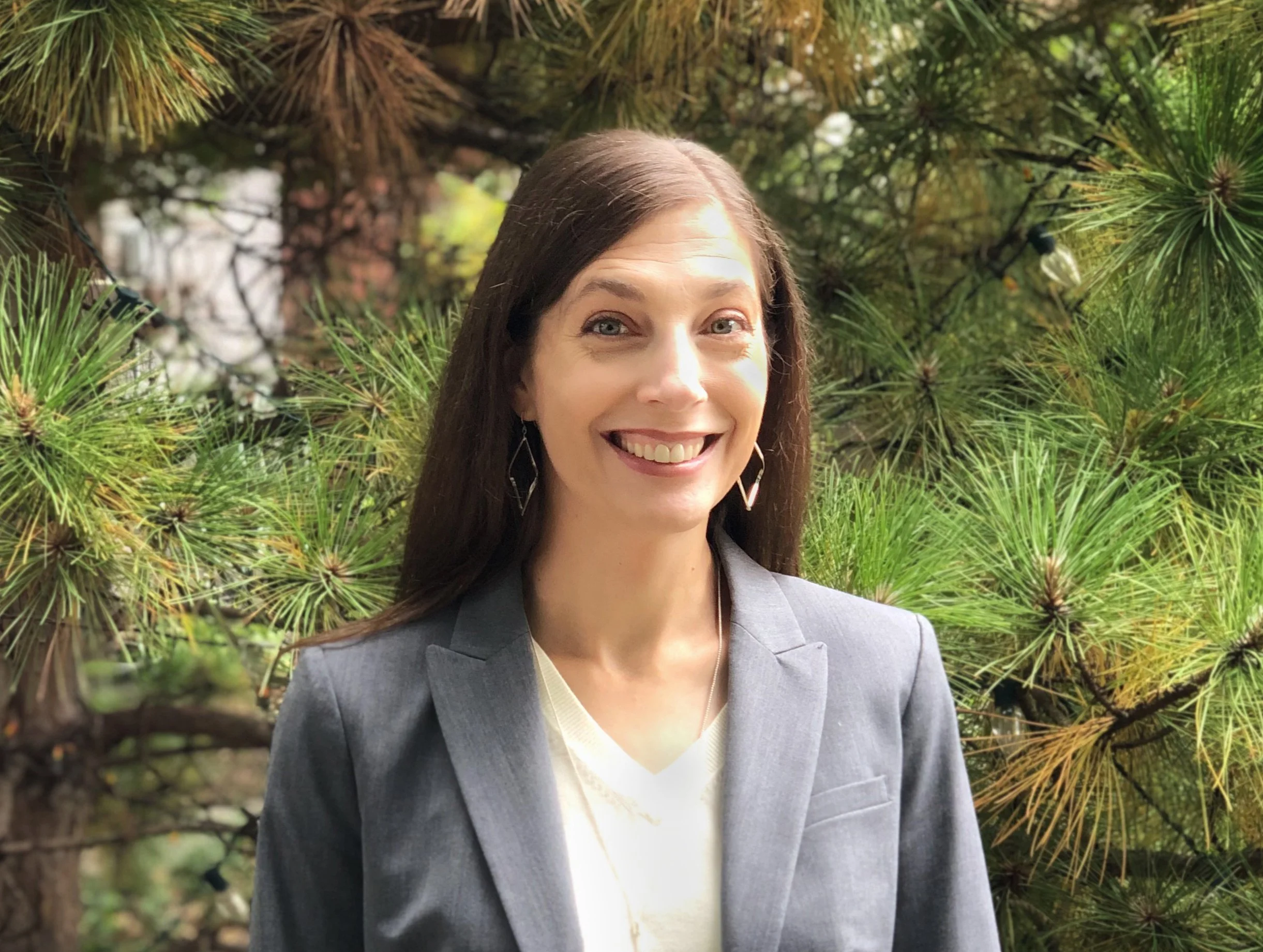 A woman, Susan Olmsted, AIA, ASLA, LEED AP BD+C with long brown hair smiling, wearing earrings and a gray blazer, outdoors in front of green pine trees.