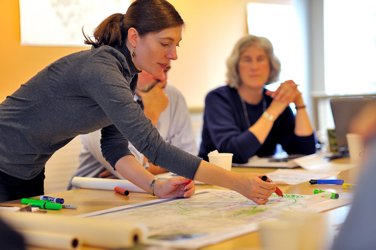 A woman in a gray top is leaning over a table, drawing or writing with colorful markers on a large sheet of paper. Two older women are seated nearby, observing. The table is scattered with markers, papers, and cups.