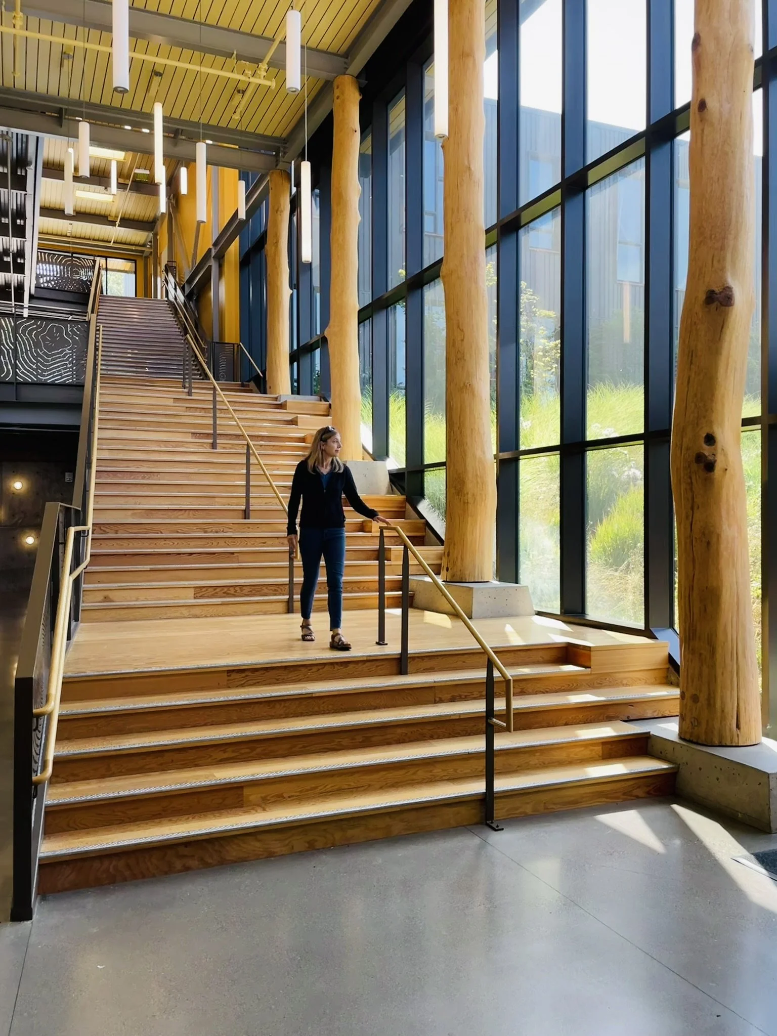 Interior 'gallery' at Blakely Elementary School with wooden stairs leading upward, large glass windows, wooden support beams, and contemporary hanging light fixtures. A woman is walking down the stairs.