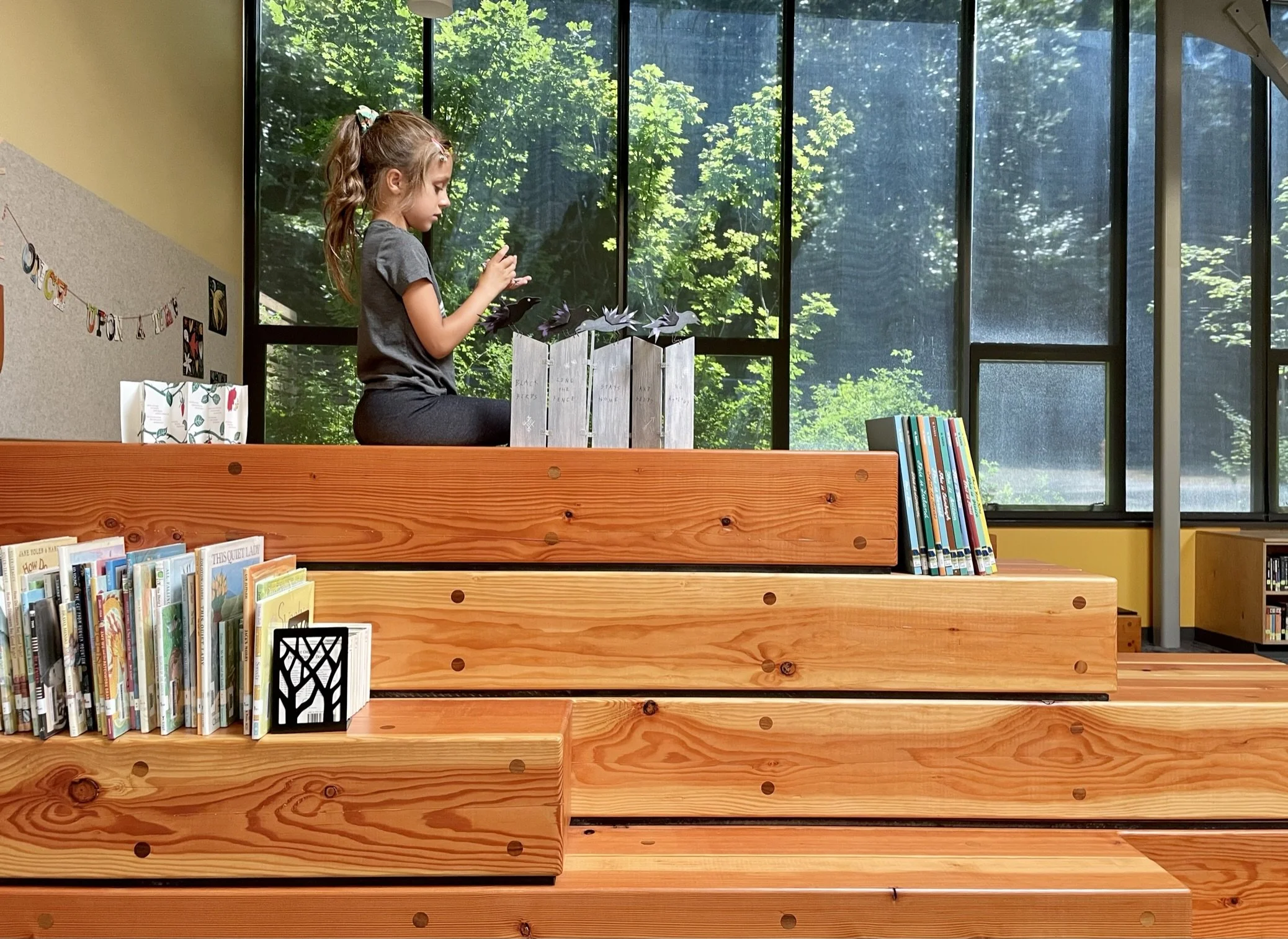 A young girl reading a book inside the Blakely Elementary School library with large windows showing green trees outside. She is sitting on a sculptural wooden surface, surrounded by books and decorations.