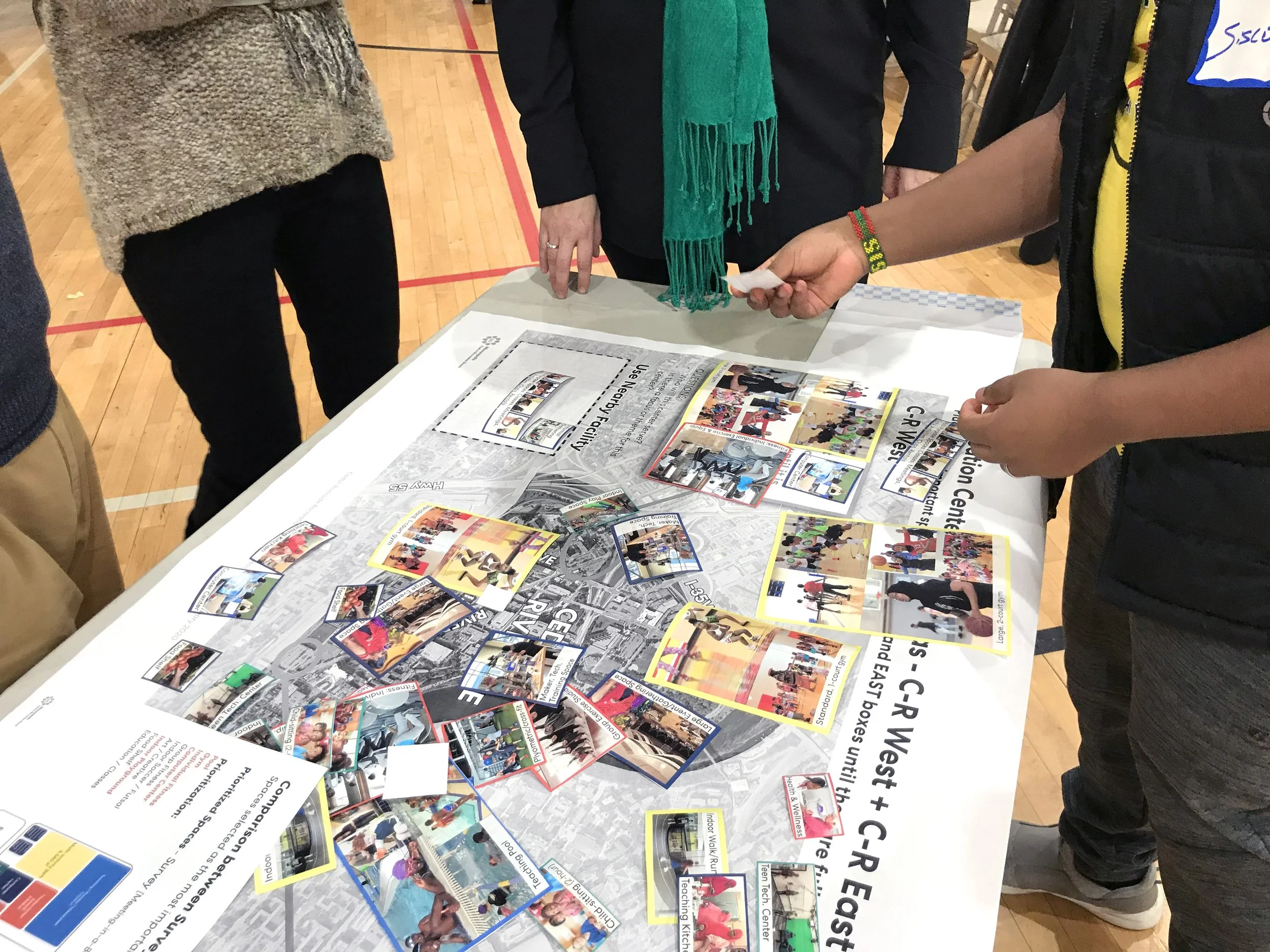 Table with colorful cards and informational flyers about a community event, with people gathered around, partly visible, inside a gymnasium.