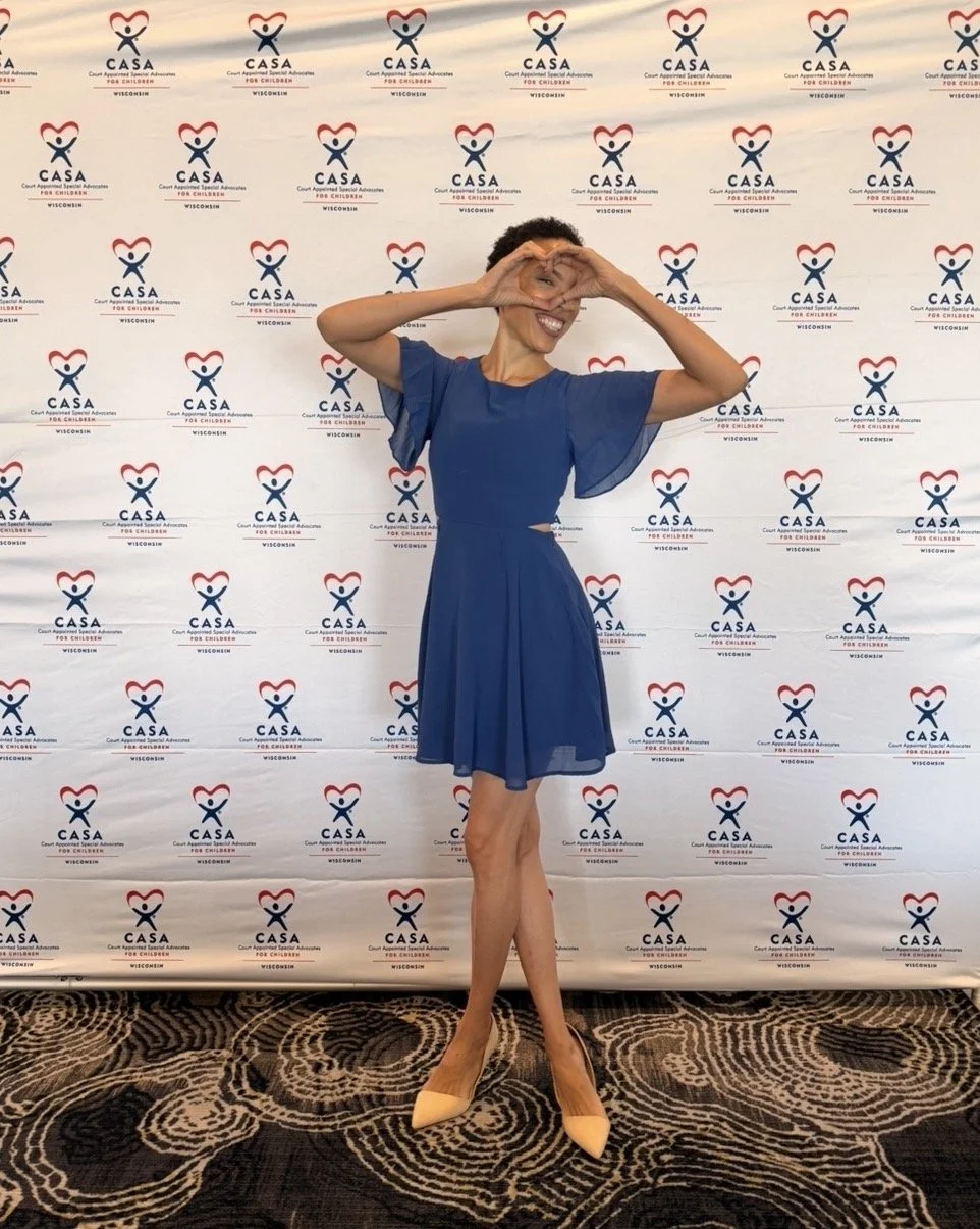 MelRo making a heart shape with her hands in front of a backdrop with the logo of CASA for Children, featuring a stylized heart and figure, at an event in Wisconsin. She is wearing a blue dress and beige high heels, smiling and posing.