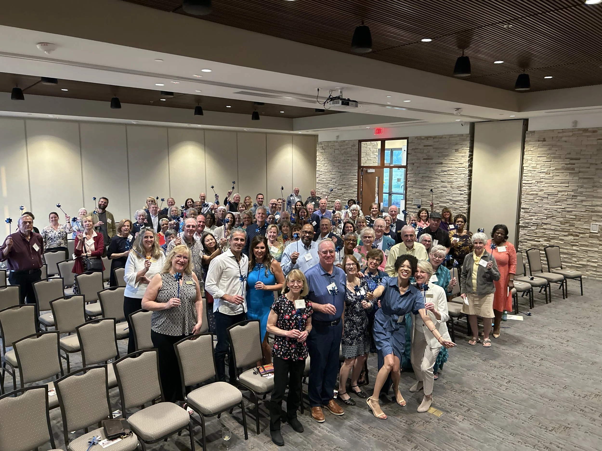 A large group of people gathered in a conference room, many holding blue and white pinwheels, smiling for the photo.