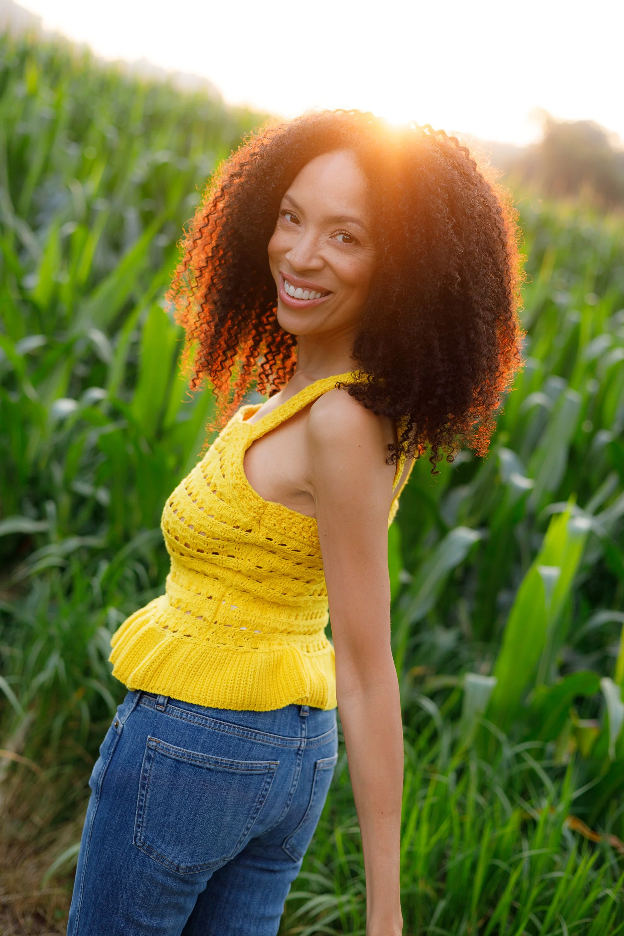 MelissaRoshan smiling with curly hair wearing a yellow crochet top and jeans standing in a green field at sunset.
