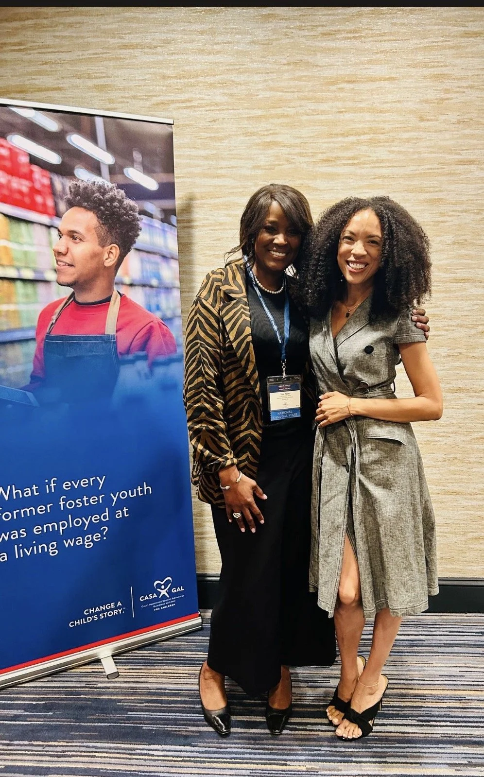 Two women standing together and smiling in front of a poster at an event. The woman on the left is wearing a black dress with a tiger-stripe blazer, and the woman on the right is wearing a gray dress with a tie at the waist. They are in a conference room with a textured beige wall and striped carpet.